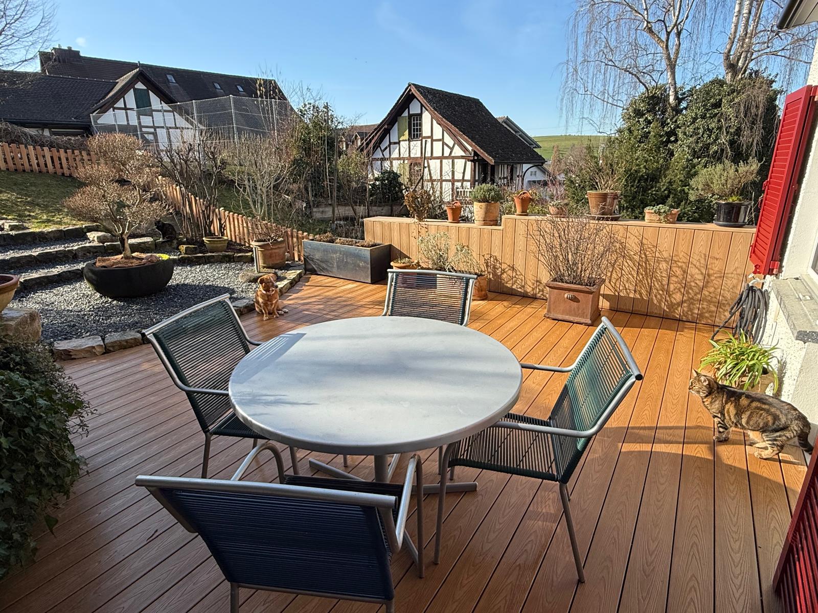 Holzdeck-Sitzplatz mit Bonsai-Schale und rundem Tisch, Riegelhaus im Hintergrund, weiches Tageslicht